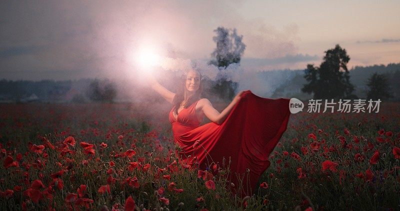 Portrait of pretty brunette girl in a red dress dancing with red burning signal flare in poppy field